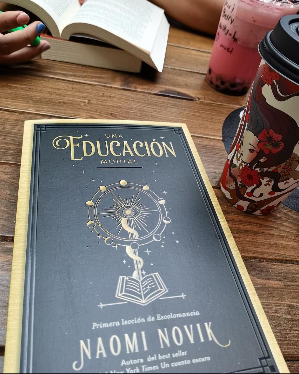 Foto de un libro de ciencia ficción, llamado 'Una educación mortal', junto con un vaso de café en un grupo de lectura.